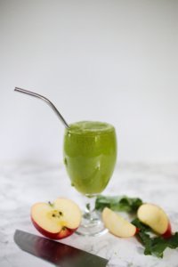 green pineapple apple smoothie in glass. Apples and knife on counter. Metal straw in glass.