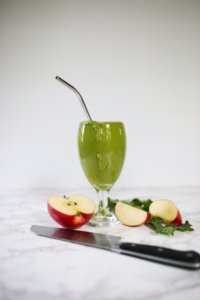 green pineapple apple smoothie in glass. Apples and knife on counter. Metal straw in glass.