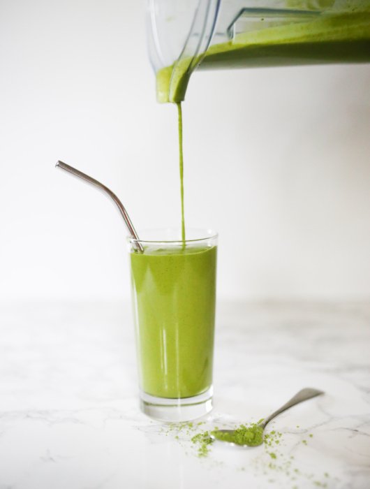 Green Matcha Maca smoothie in tall glass with metal straw, smoothie being poured from vitamix blender