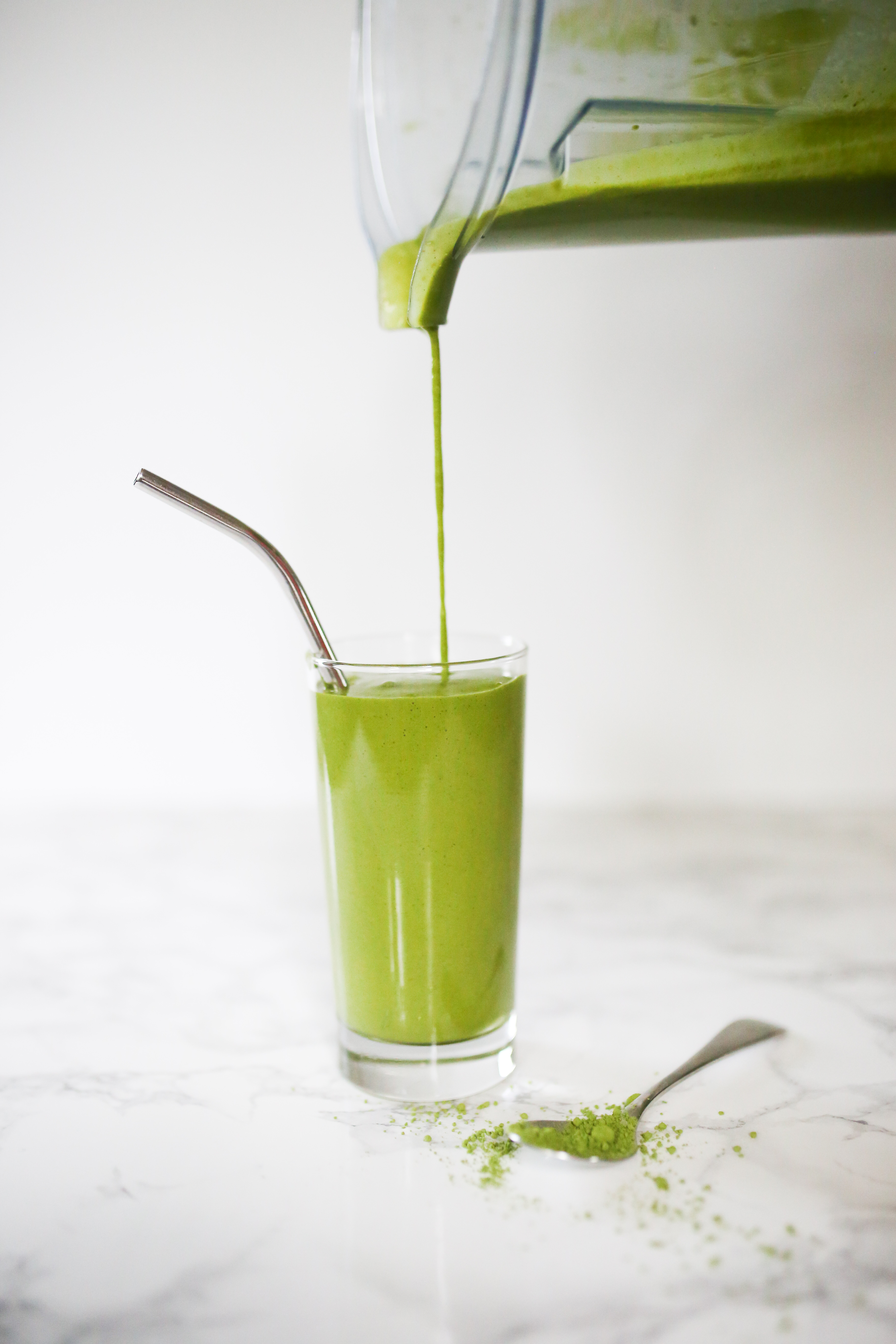 Green Matcha smoothie in tall glass with metal straw, smoothie being poured from vitamix blender